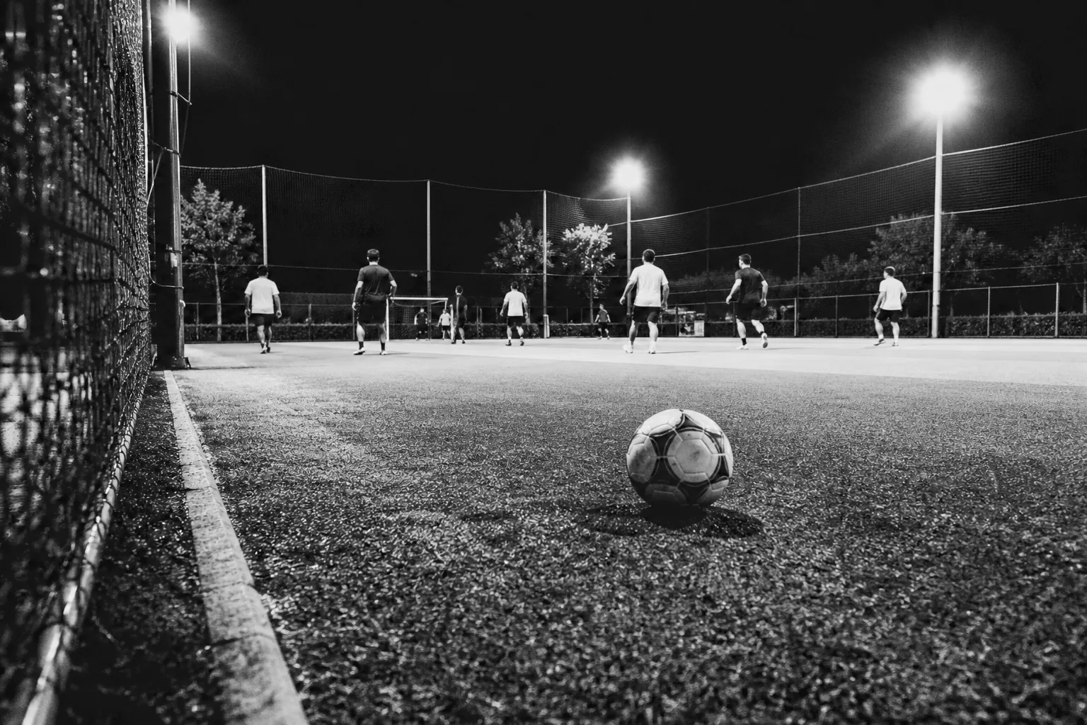 Cancha de fútbol sintética iluminada de noche, con jugadores al fondo y un balón en primer plano